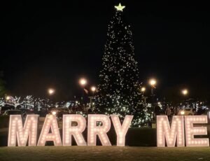 Marquee letters spelling out. "Marry Me" for engagement in front of Christmas tree